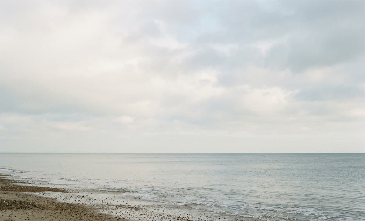 A calm grey sea meeting a shingle beach under a wide overcast sky